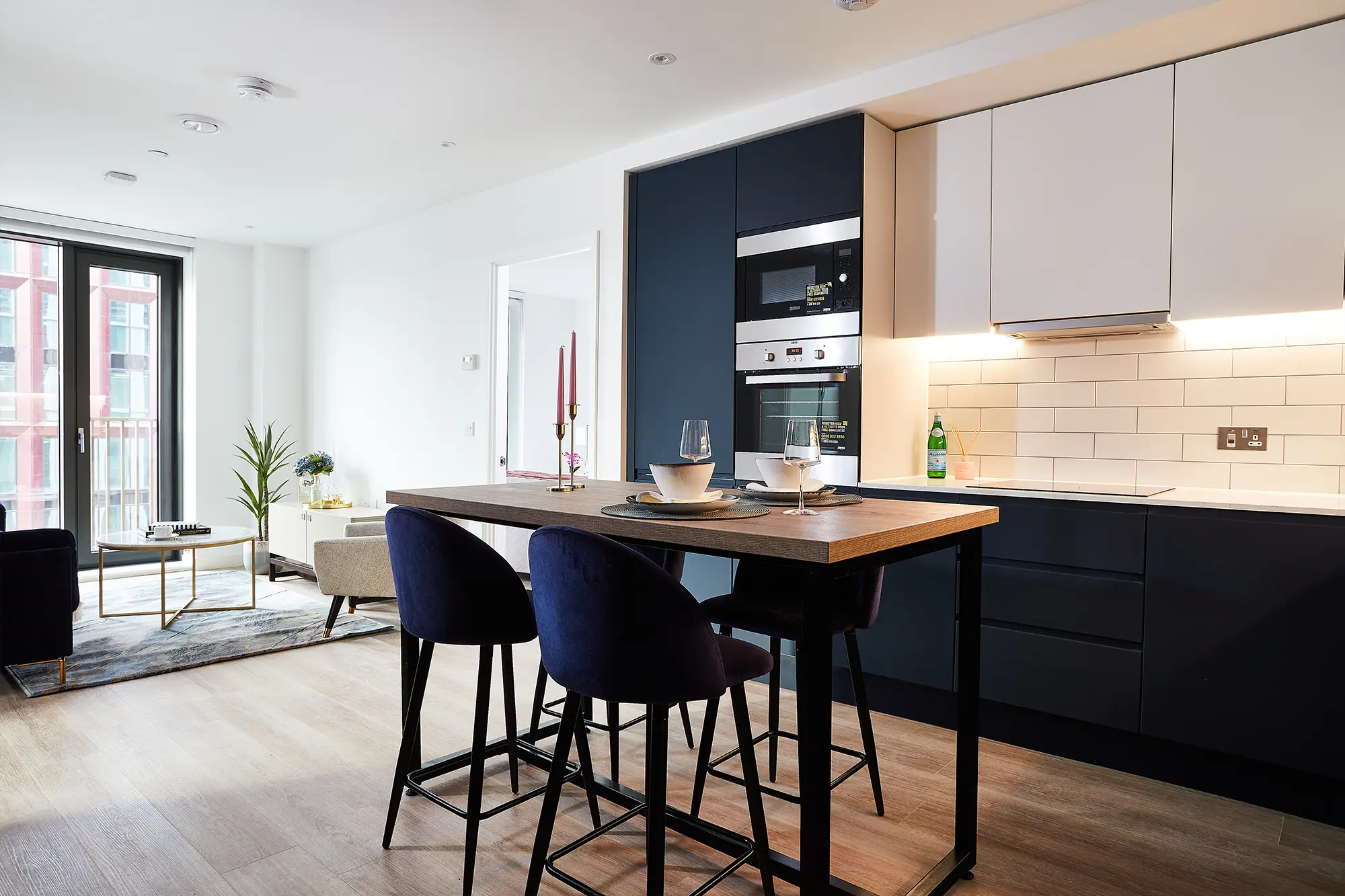 Open-plan kitchen and living area in a Vita Living Circle Square North apartment, with a navy fitted kitchen, breakfast bar seating and floor-to-ceiling doors onto a balcony