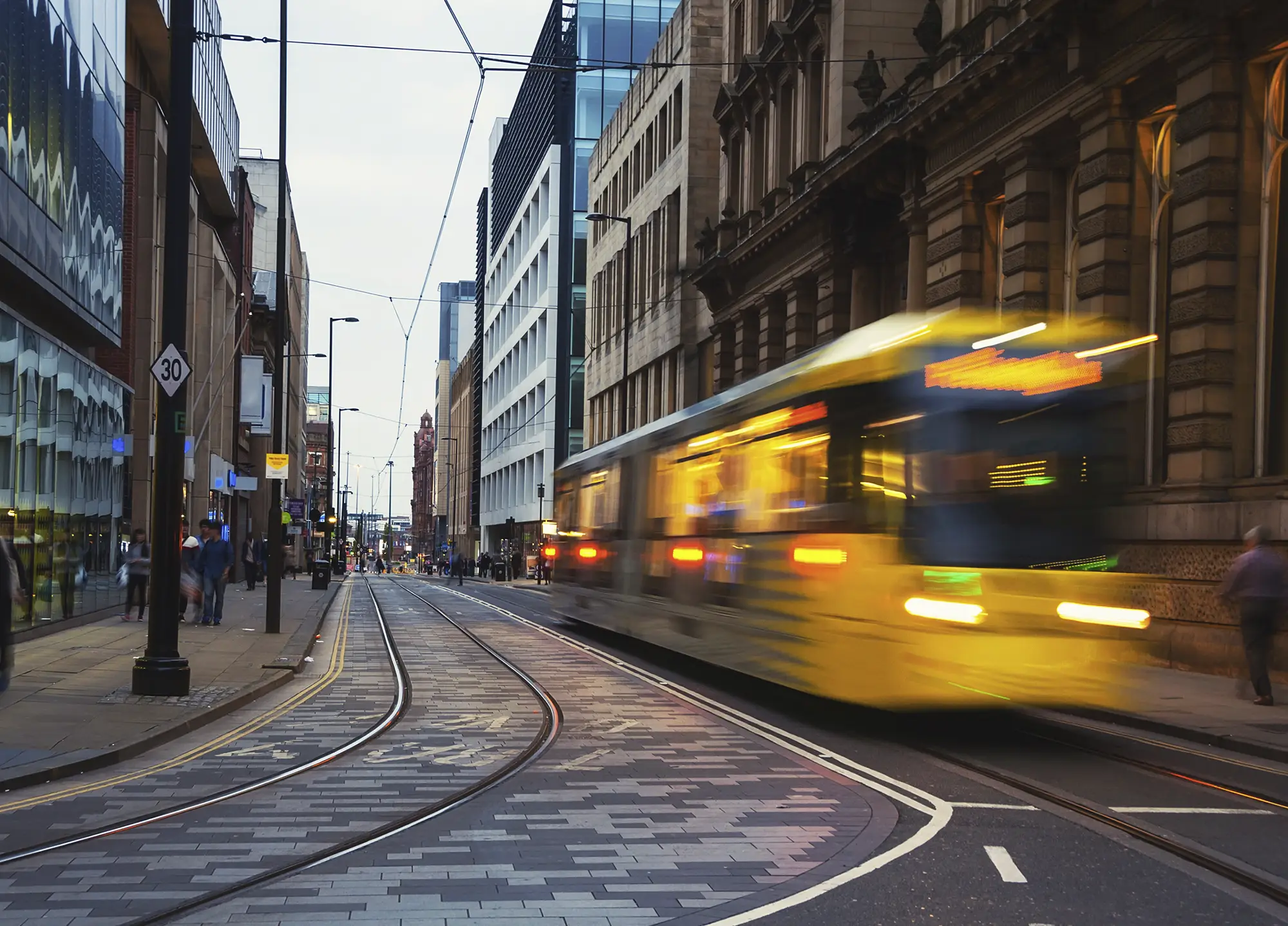 A yellow Metrolink tram passing through Manchester city centre at dusk